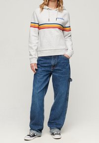 Szara bluza z kapturem z czerwonymi, żółtymi i niebieskimi paskami oraz przednią kieszenią, zestawiona z luźnymi niebieskimi dżinsami i czarnymi tenisówkami. Prosty, codzienny design.