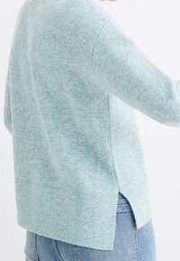 Light teal sweater made from soft fabric, featuring a relaxed fit, ribbed hem, and side slits, paired with light-wash jeans.