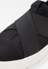 Baskets noires à enfiler, dotées de larges bandes élastiques croisées et d'une semelle blanche, conçues pour un enfilage facile et un maintien sûr.