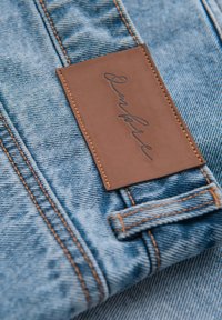 Primo piano di tessuto denim blu con cuciture arancioni e un'etichetta in pelle marrone cucita sulla vita.