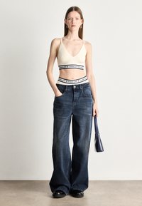 Krémová rebrovaná braletka s hlbokým výstrihom do V, spárovaná s tmavomodrými širokými nohavicami s gombíkovým zapínaním a prednými vreckami. Drží malú tmavú kabelku.