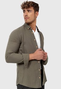 Camisa de color verde oliva hecha de algodón, con cierre de botones en la parte delantera, mangas largas y bajo curvado. Camiseta blanca debajo.