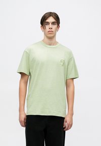 Camiseta de algodón verde claro con un corte holgado, cuello redondo y un sutil logotipo bordado en el pecho. Lleva unos pantalones negros.