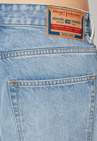 Un jean en denim bleu clair présente un patch en cuir marron avec des accents bleus et rouges, cousu avec un fil orange, mettant en valeur la poche arrière.