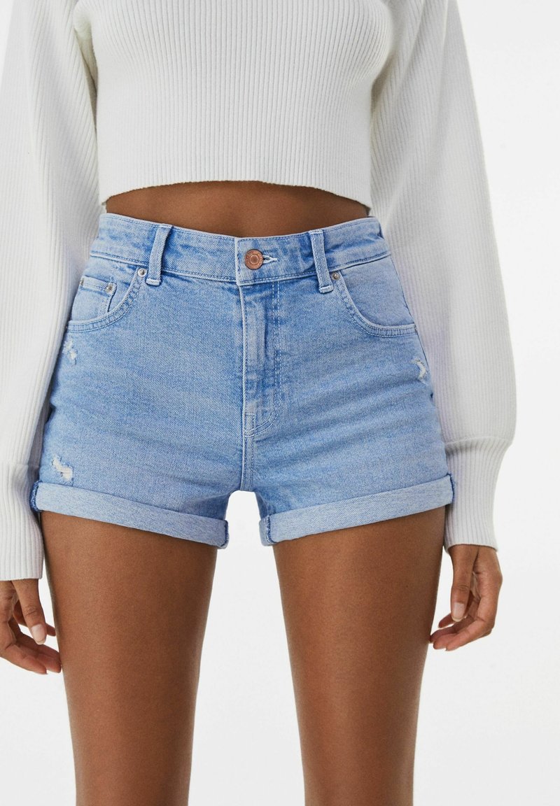 Ljust blå denimshorts med uppvikta muddar, med en femfickorsdesign och lätt nött detaljering. Knappar och sömmar är i kontrasterande färg.