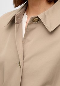 Zbliżenie na kolor beżowy kołnierz płaszcza i dwa guziki, noszonego na białej koszuli, ukazujące detale haftu i fakturę tkaniny.