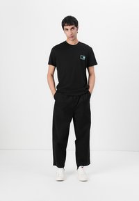 T-shirt en coton noir avec un graphisme turquoise sur la poitrine gauche, assorti à un pantalon noir ample et des baskets blanches. Look décontracté et design.
