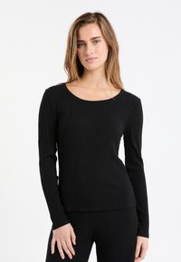 Kobieta z jasnobrązowymi włosami, ubrana w dopasowany czarny top z prążkowanym wzorem z długim rękawem oraz pasujące czarne spodnie prążkowane, na tle białym.