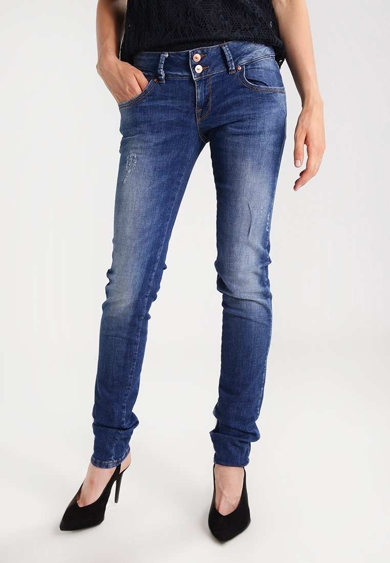 Femme portant un jean bleu slim légèrement usé et des chaussures noires à talons hauts, debout avec une main dans la poche.