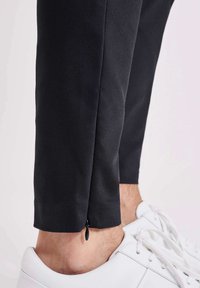 Schwarze schmal zulaufende Hose mit glatter Textur, ausgestattet mit einem Reißverschlussdetail am Knöchel, kombiniert mit weißen Sneakern und einer Nahaufnahme des Saums.