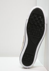 Zapatilla de lona color beige con una suela de goma negra texturizada, detalles en blanco y un patrón clásico de diamantes. Presenta un logo embellecido en la suela.