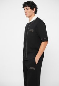 Homem em camisa de botão de manga curta e conjunto de calças pretas com logótipos "Armani Exchange N.Y.", com a mão no bolso, voltado para a esquerda em fundo branco.