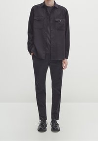 Uomo che indossa una camicia nera con bottoni e tasche a soffietto, pantaloni abbinati e scarpe stringate in pelle nera, in piedi contro uno sfondo chiaro e semplice.