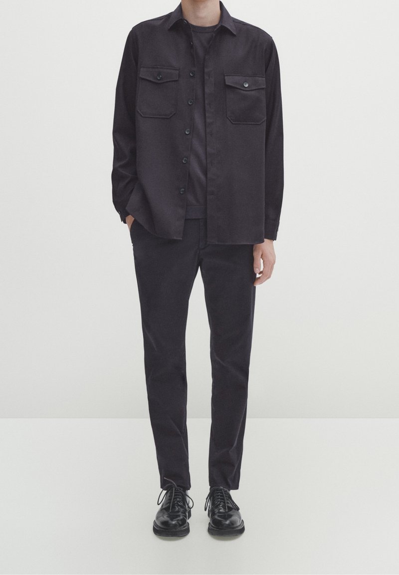 Uomo che indossa una camicia nera con bottoni e tasche a soffietto, pantaloni abbinati e scarpe stringate in pelle nera, in piedi contro uno sfondo chiaro e semplice.