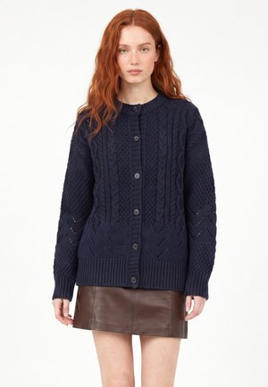 Kvinde med langt rødt hår iført en mørkeblå kabelstrikket cardigan med knapper og et mørkebrunt lædernederdel mod en hvid baggrund.
