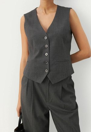 Gilet sans manches gris foncé boutonné associé à un pantalon plissé taille haute assorti, personne tenant un sac à main noir.