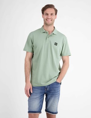 Man die lacht, draagt een lichtgroen poloshirt met korte mouwen en blauwe spijkershorts, staat met één hand in zijn zak tegen een effen witte achtergrond.