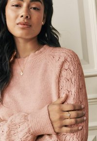 Gaiši rozā adīts džemperis ar ribotu apkakli un mežģīņu raksta piedurknēm. Izskatu izceļ zelta kaklarota un gredzens. Redzama gluda tekstūra.