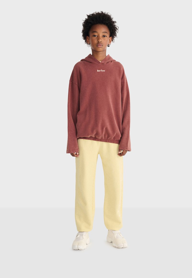 Young person standing, wearing a rust-colored oversized hoodie, light yellow pants, and white chunky sneakers, against a plain light background.