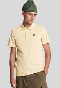 Geel poloshirt met korte mouwen, twee knoopsluitingen en zwart geborduurd logo op de linkerkant van de borst. Gedragen met kaki shorts en een groene gebreide muts.