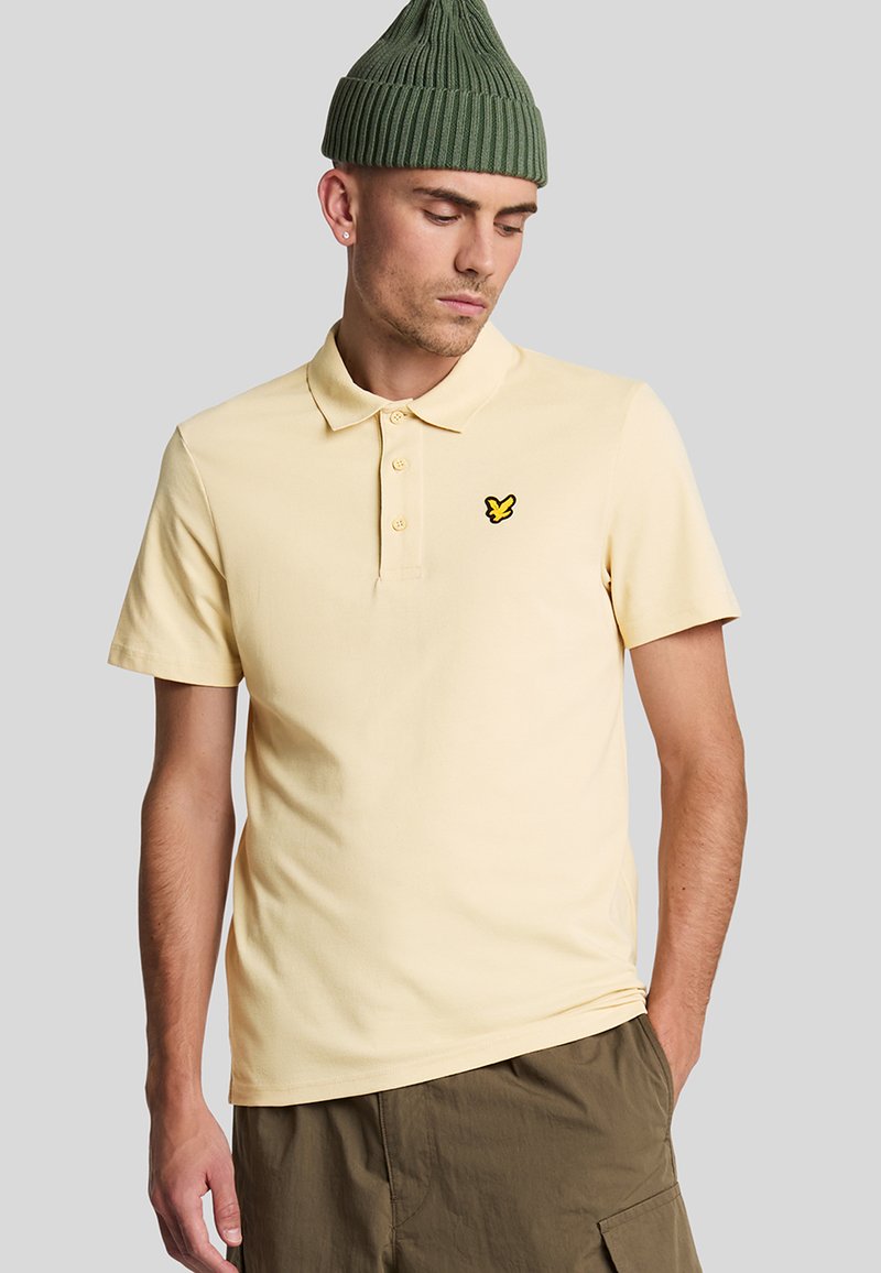 Geel poloshirt met korte mouwen, twee knoopsluitingen en zwart geborduurd logo op de linkerkant van de borst. Gedragen met kaki shorts en een groene gebreide muts.