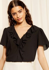 Tricou negru cu mâneci scurte, cu decolteu în V și detalii cu volane pe partea din față, realizat dintr-un material texturat cu un model subtil.