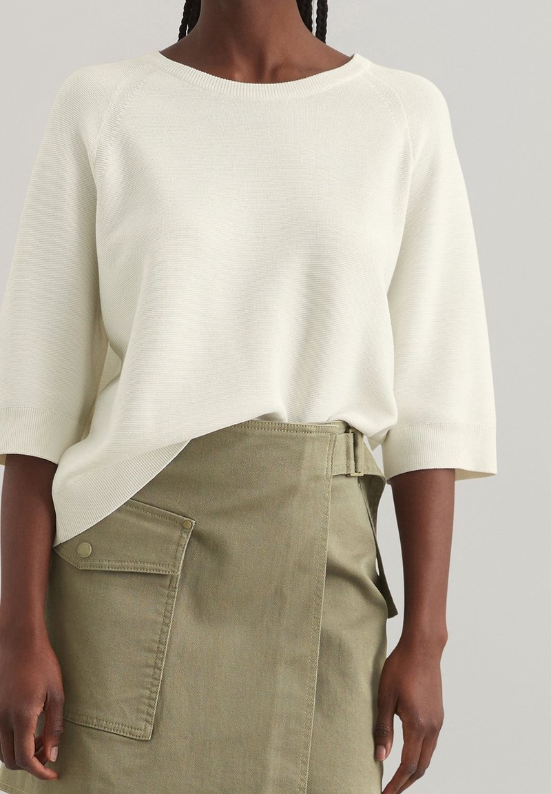 Femme portant un haut en maille blanc à manches courtes rentré dans une jupe portefeuille vert olive avec une poche latérale et un détail ceinture.