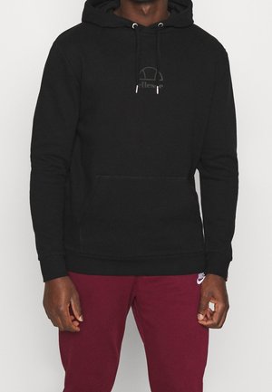Mand iført sort hættetrøje med forlommer og bordeaux Nike træningsbukser mod en ensfarvet baggrund.