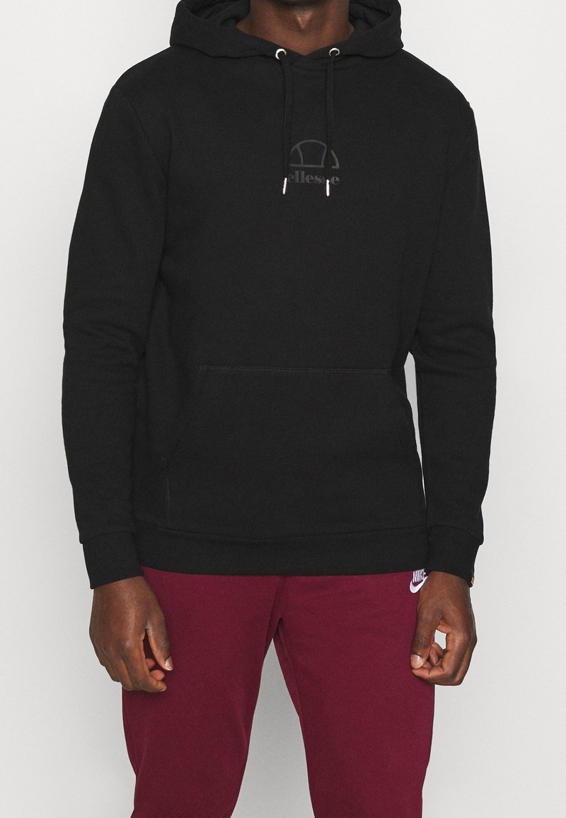 Homme portant un sweat à capuche noir avec poche avant et un pantalon de survêtement Nike bordeaux, sur un fond uni.