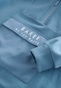 Maglione blu con tessuto strutturato e una tasca frontale liscia sovrapposta. Presenta una cerniera argento e un logo bianco del marchio sulla tasca.