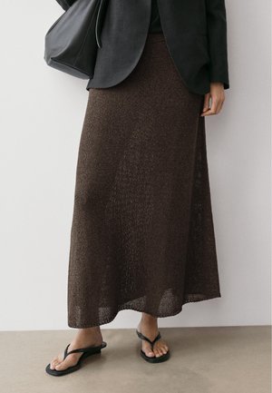 Onderste helft van een persoon die een lange bruine gebreide rok, een zwart blazer, zwarte slippers draagt en een zwarte tas bij zich draagt.