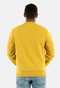 Sudadera amarilla con cuello redondo y puños acanalados. La parte trasera cuenta con un diseño minimalista, combinada con vaqueros de color azul claro.