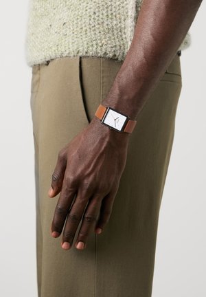 Mørkhudet arm med firkantet ur med band i lysebrunt læder, mod baggrund af khakibukser og en lyse tekstureret sweater.