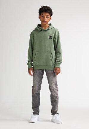 Sudadera verde con capucha y bolsillo delantero, mangas largas y parche con logo circular negro. Combina con jeans grises y zapatillas blancas.