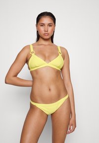 Bikini amarelo com riscas, apresentando um top triangular com detalhes em metal redondos e cuecas de corte baixo a condizer. Textura suave, design minimalista.
