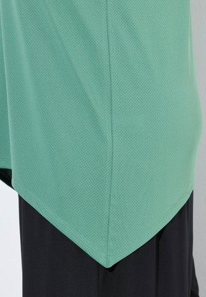 Grün perforiertes Sportoberteil mit einem stylischen Saum, kombiniert mit schwarzen Hosen. Fokus auf atmungsaktivem Material und schrägen Design.