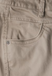Pantalon beige avec une poche avant dotée de coutures visibles et d'un détail en rivet métallique. Le tissu a une texture lisse.