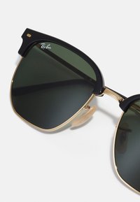 Black and gold sunglasses with green lenses. Features a metal frame, plastic accents, and a prominent nose pad. Ray-Ban logo visible on frame.