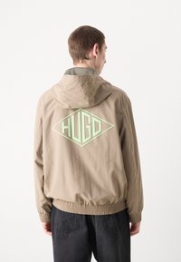 Lys beige hettegenser med elastiske mansjetter og kant, med en stor mintgrønn "HUGO" logo i diamantform på baksiden.