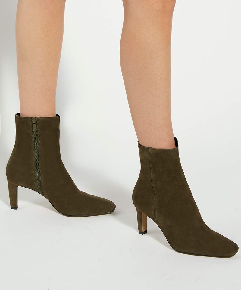 Jambes portant des bottines en daim vert olive avec des fermetures éclair latérales et des talons blocs de hauteur moyenne, posées sur une surface blanche.