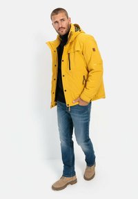 Gul isoleret jakke med hætte, frontlynlås og flere lommer, kombineret med blå denimjeans og beige støvler.