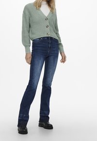 Kvinna klädd i en ljusgrön grovstickad kofta, vit polotröja, mörkblå bootcut-jeans och svarta grova stövlar som står mot en vit bakgrund.