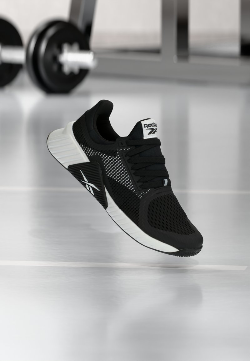 Ασπρόμαυρο αθλητικό παπούτσι Reebok που αιωρείται πάνω από ένα λαμπερό δάπεδο γυμναστηρίου με θαμπά βάρη γυμναστηρίου στο φόν το.