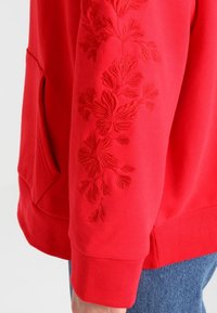 Sudadera roja con un diseño de bordado floral texturizado en la manga izquierda, que presenta mangas largas y un corte holgado.