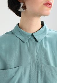 Una camisa de color verde agua claro, con cuello y dos bolsillos delanteros, hecha de tela suave. Presenta un cierre con botones y una textura lisa y uniforme.