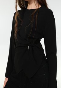Fekete hosszú ujjú felső, csavart derékrésszel, finom cipzár részlettel és sima, rugalmas anyagtextúrával.