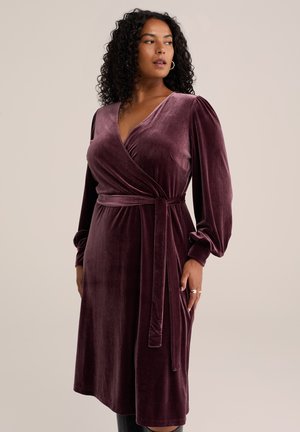 Femme portant une robe portefeuille en velours violet profond à manches longues avec une ceinture à la taille, regardant sur le côté sur un fond beige uni.