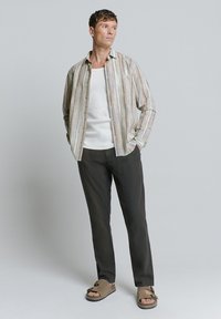 Gestreepte button-up shirt in beige en pastelkleuren, witte tanktop, donkere broeken en bruine slippers met een platte zool. Eenvoudige, ontspannen pasvorm.