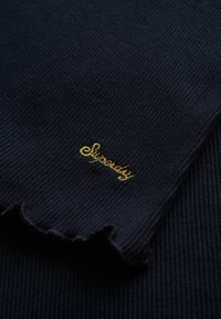 Ribbede mørkeblå stof med en bølget kant, prydet med guld broderet tekst "Superdry" på forsiden, der fremhæver branddetaljen.