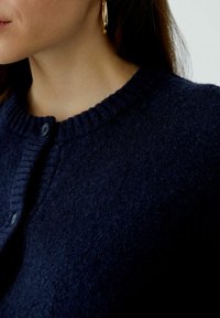 Dunkelblauer, strukturiert gestrickter Cardigan mit ribbeltem Rundhalsausschnitt, Knopfverschluss und weichem Wollmaterial. Nahaufnahme des Oberteils.